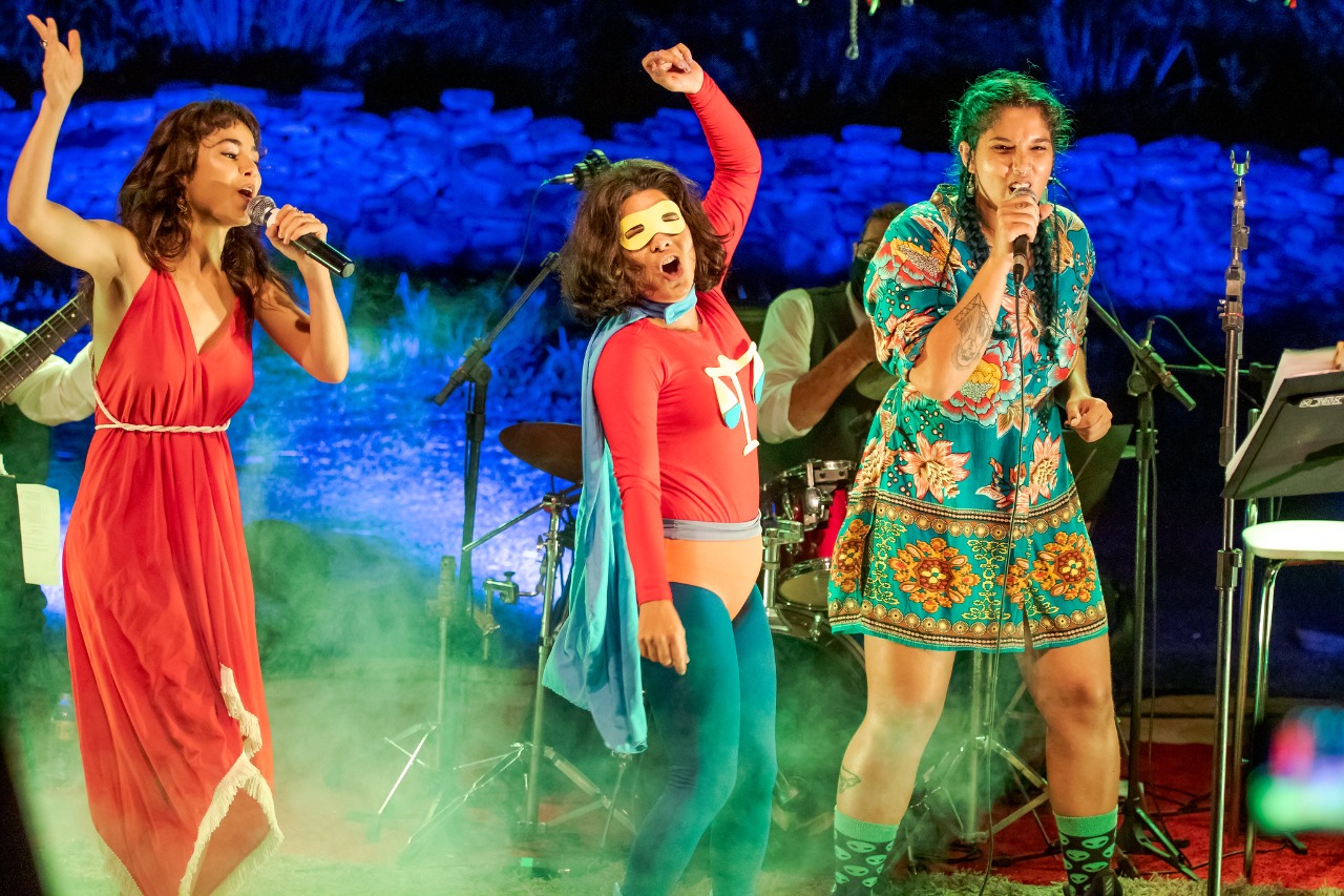 #PraCegoVer Em destaque, na foto, estão três mulheres posicionadas durante uma apresentação cultural: a cantora Flaira Ferro, a personagem que representa o bloco, e mais uma artista.
